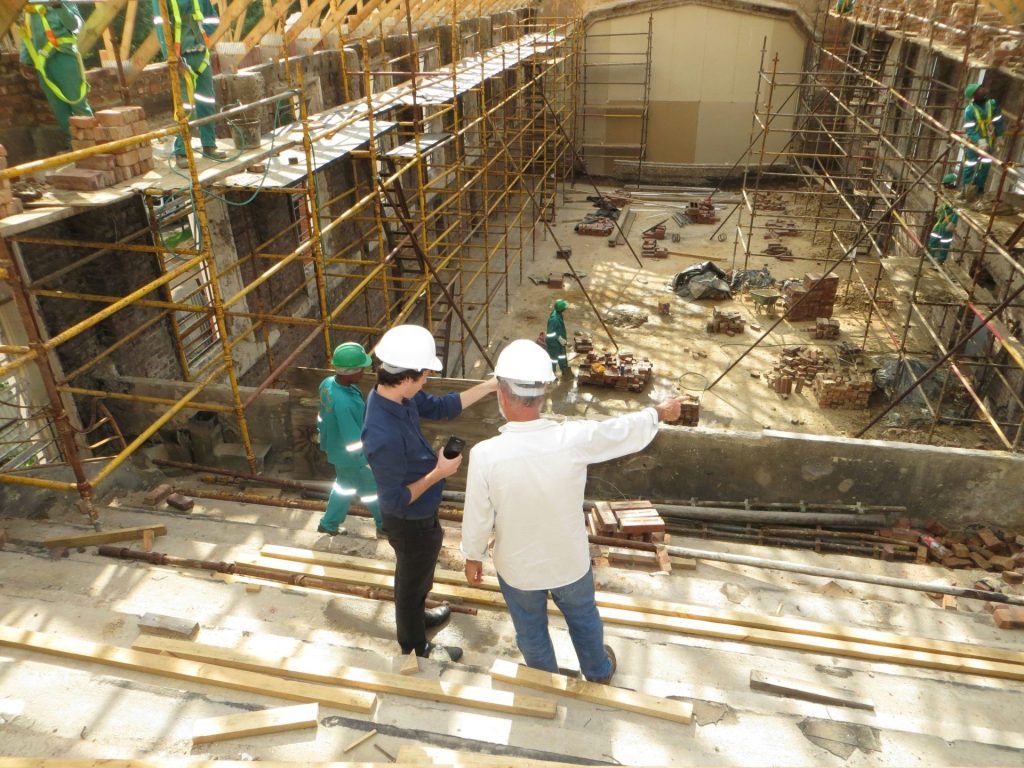 Zwei Männer mit Schutzhelmen zeigen auf eine Baustelle mit Gerüst.